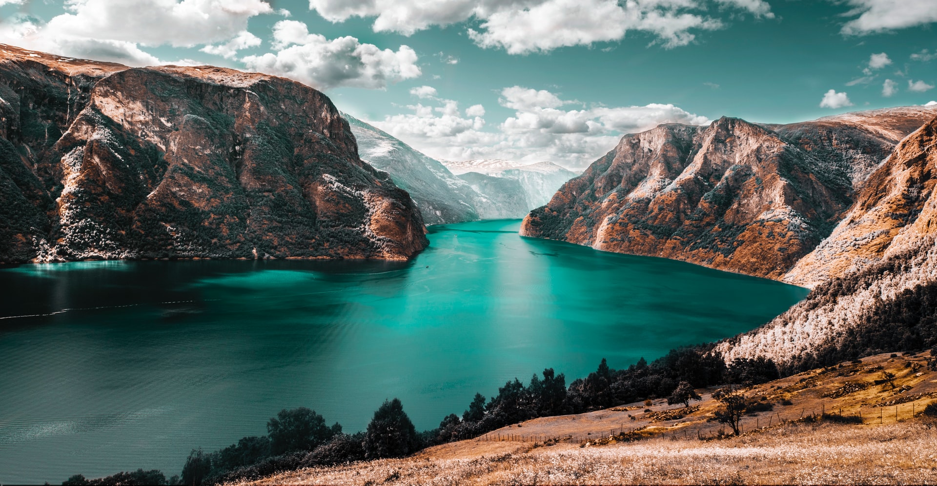 Turquoise fjord flanked by steep mountains with sparse vegetation under a partly cloudy sky.
