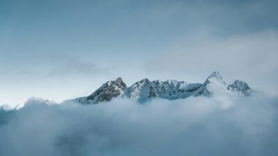 Snow-covered mountain peaks surrounded by fog under a soft blue sky.