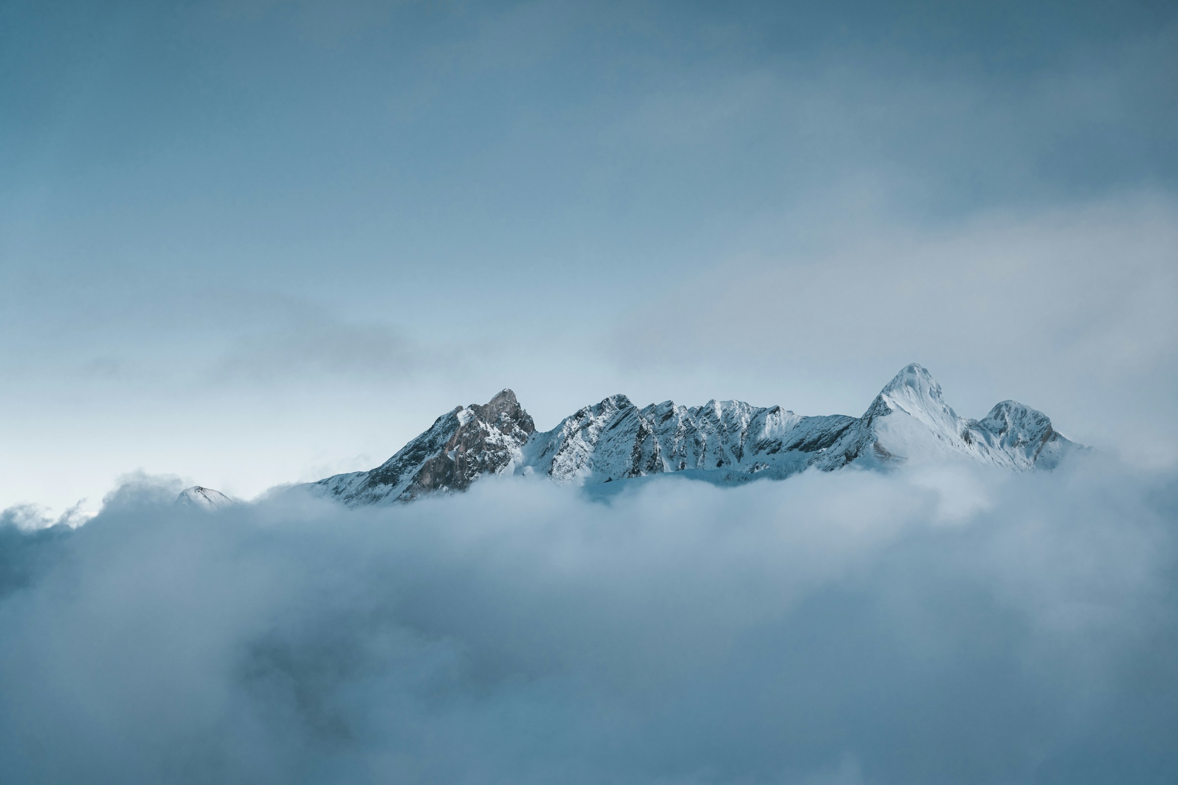 Snow-covered mountain peaks surrounded by fog under a soft blue sky.