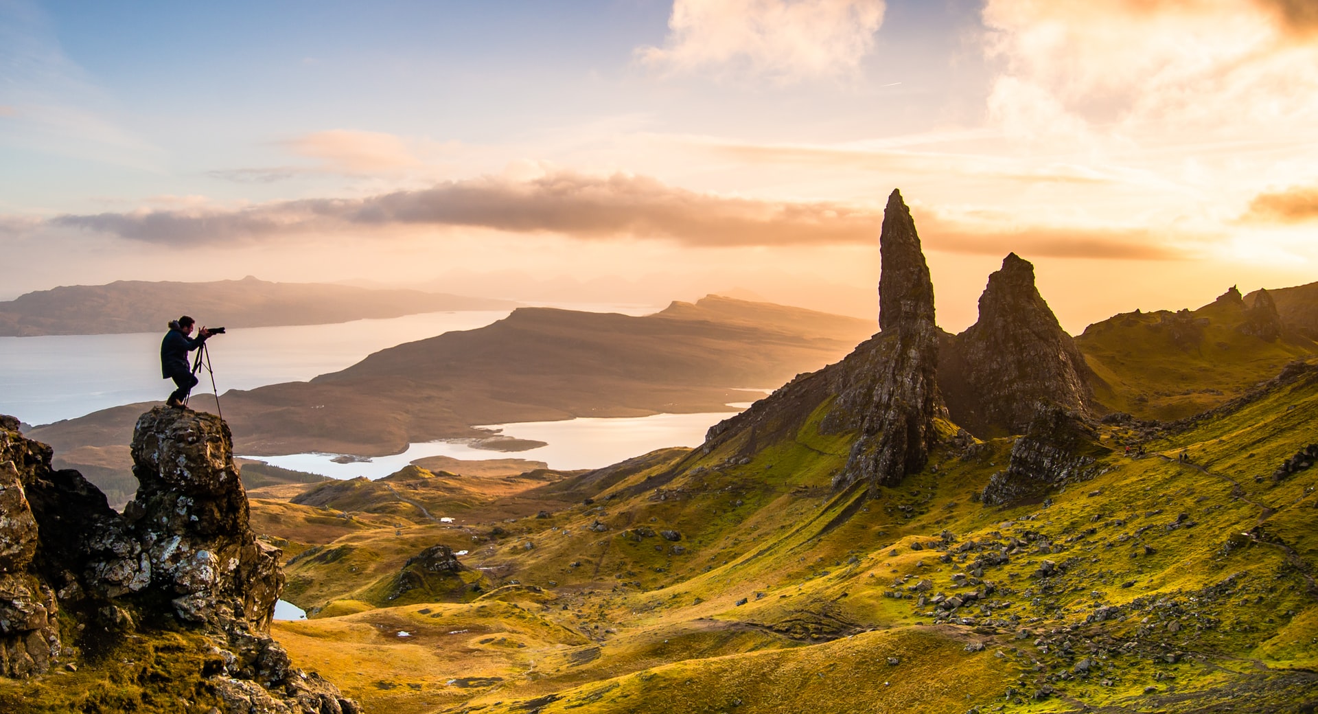 Landscape of green rolling hills and rocky formations at sunrise with water bodies in the distance.