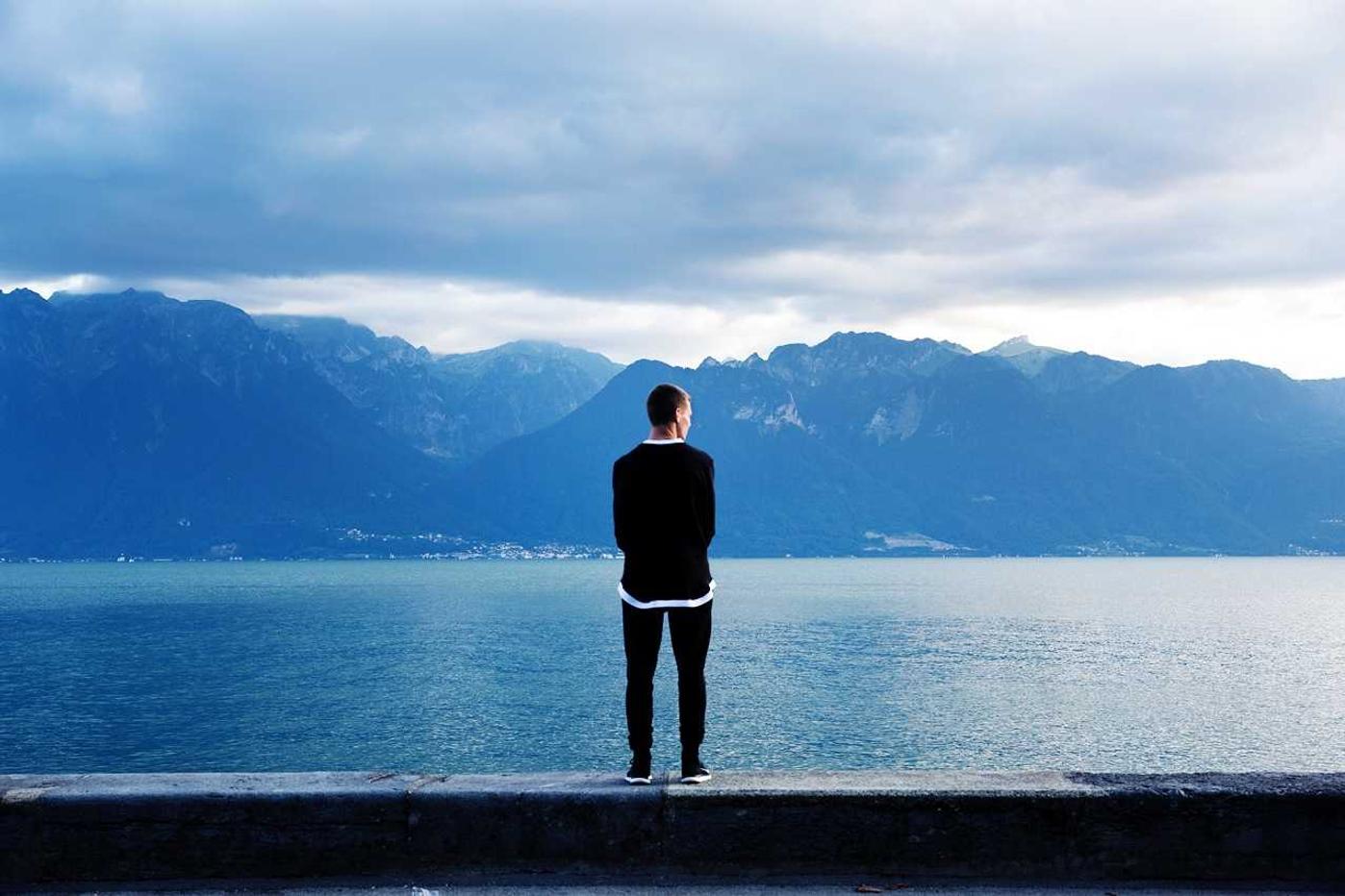 A person in black stands on a concrete ledge overlooking a calm lake surrounded by mountains.