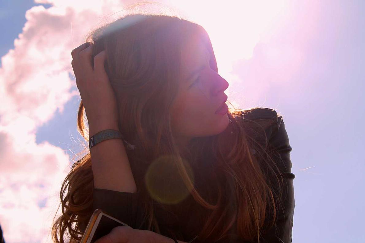 A person with long hair holds their head, with sunlight and lens flare behind them.