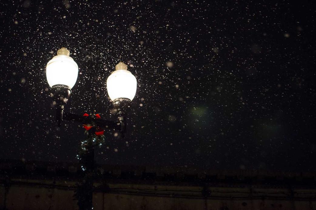 Two glowing street lamps at night with snowflakes falling around them.