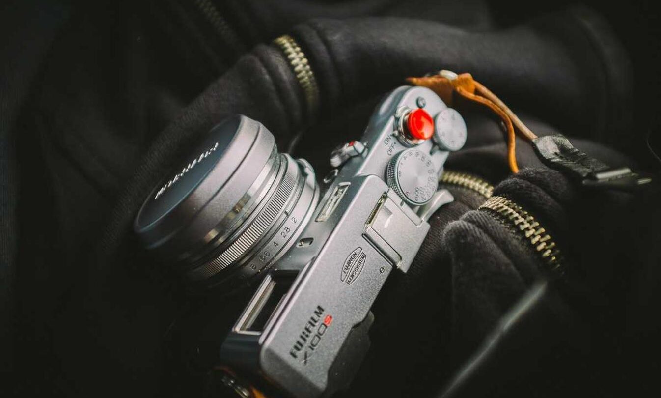 A silver vintage-style film camera with a leather strap rests on a black fabric background with visible zippers.