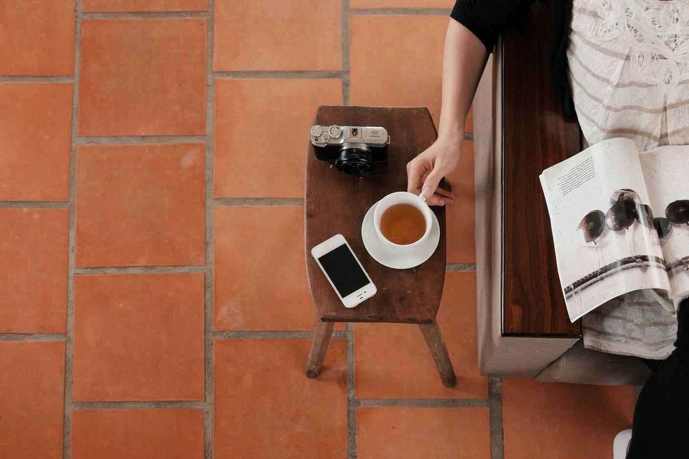 A hand reaching for a cup of coffee on a small wooden table, next to a smartphone and a camera.