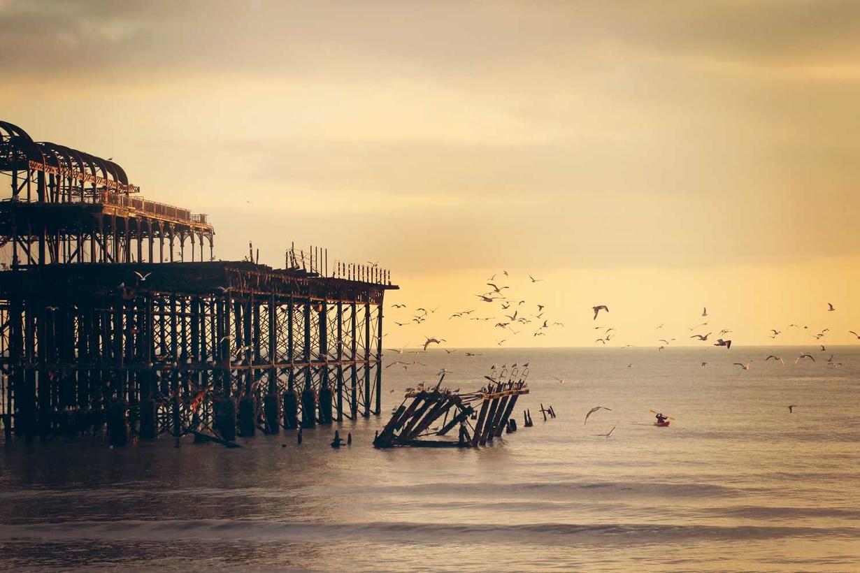 A partially collapsed pier extending into calm water during sunset, with birds flying nearby.